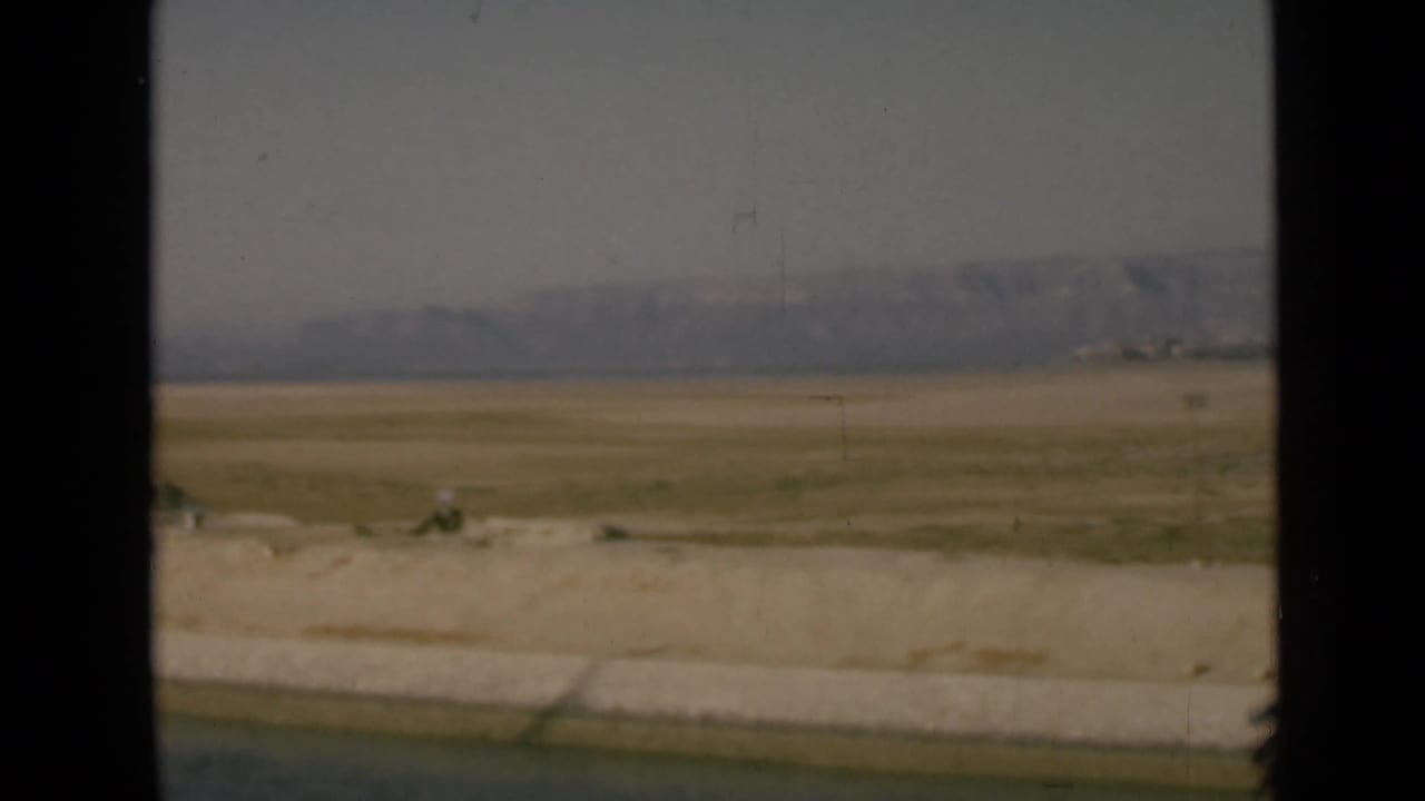 Canal-side desert view in Egypt, with a concrete edge in the foreground, flat arid land, distant hills, and visible 8mm film grain.