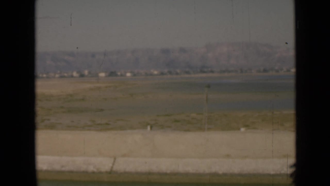 8mm home-movie frame from the Suez Canal region showing desert flats, a faint distant settlement, low hills, and foreground roadside blur.