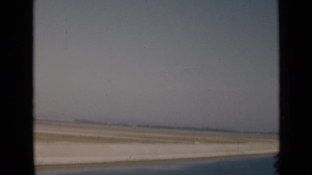 Soft 8mm frame of the Suez Canal region, showing water, sandy bank, open desert, and a distant hazy horizon.
