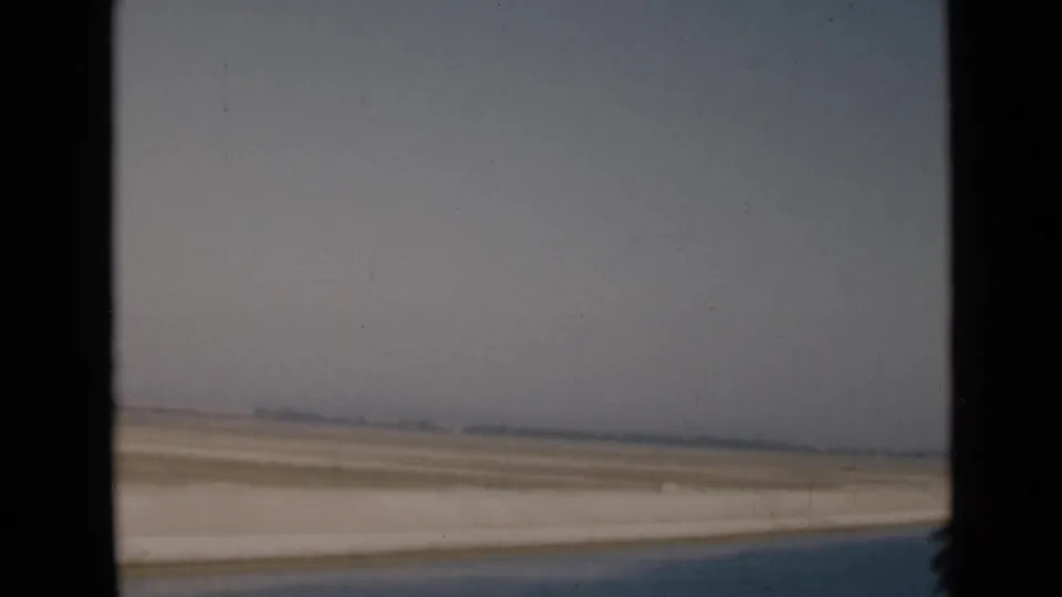 Soft 8mm frame of the Suez Canal region, showing water, sandy bank, open desert, and a distant hazy horizon.