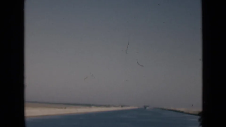 Shipboard 8mm view of the Suez Canal in Egypt, with water, sandy banks, haze, and dark vessel framing.