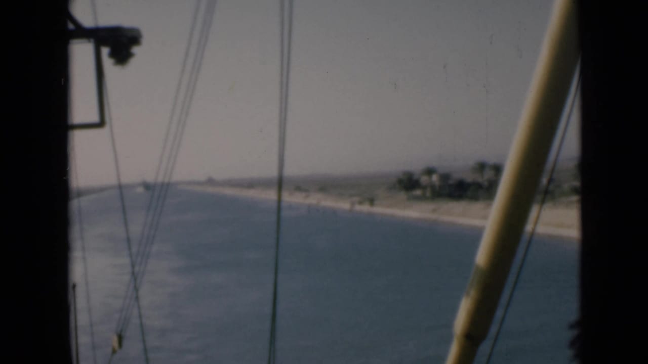 1946 shipboard view along the Suez Canal, with rigging lines, a pale boom, water, and low sandy shore.