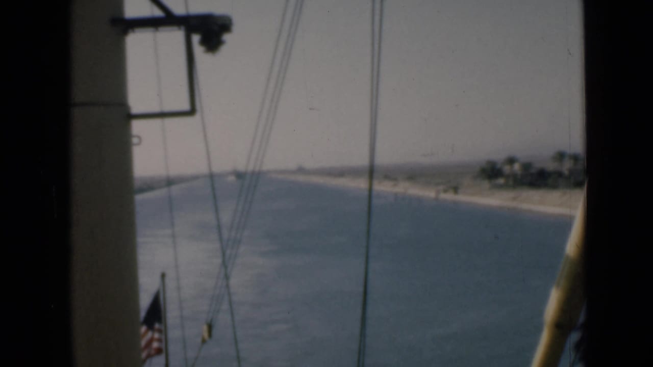 Shipboard Suez Canal view with rigging, partial American flag, blue water, sandy bank, and distant vegetation.