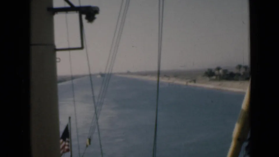 Shipboard Suez Canal view with rigging, partial American flag, blue water, sandy bank, and distant vegetation.