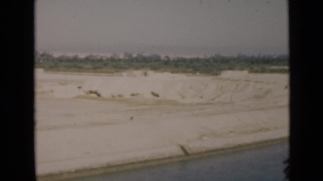 1946 Suez Canal shoreline from a moving vessel, with blue water, pale sand, and a low green horizon.