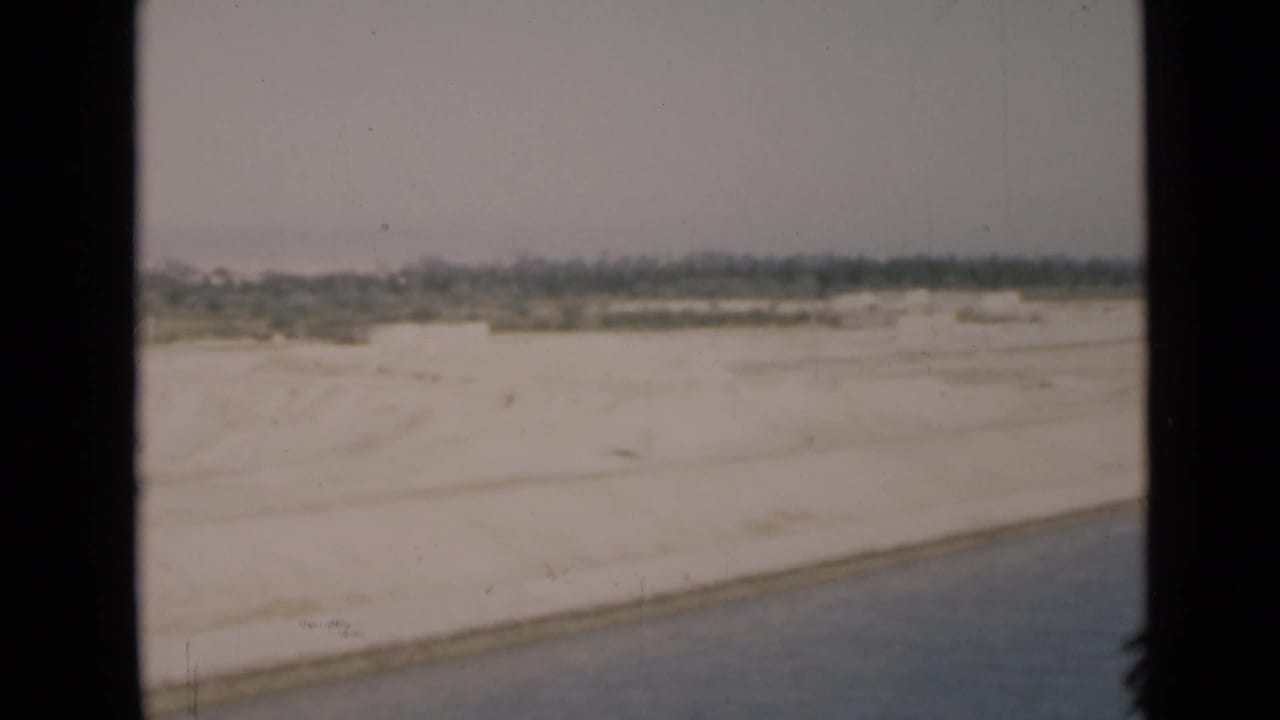 Soft 8mm frame of the Suez Canal edge, showing sand flats, water, and a low treeline in the distance.