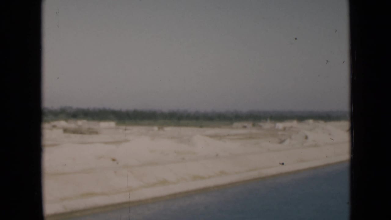1946 Suez Canal home-movie frame showing blue water, white sand, and a low green horizon through dark 8mm film borders.