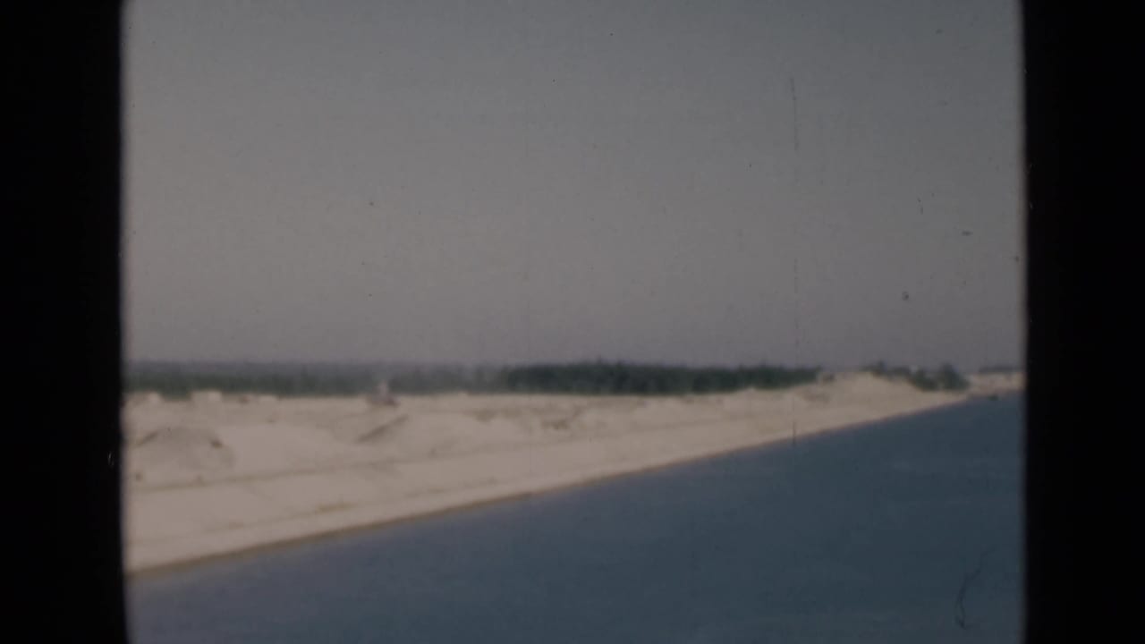 1946 Suez Canal home-movie frame with blue water, white sand, distant green shoreline, dark borders, and visible film wear.