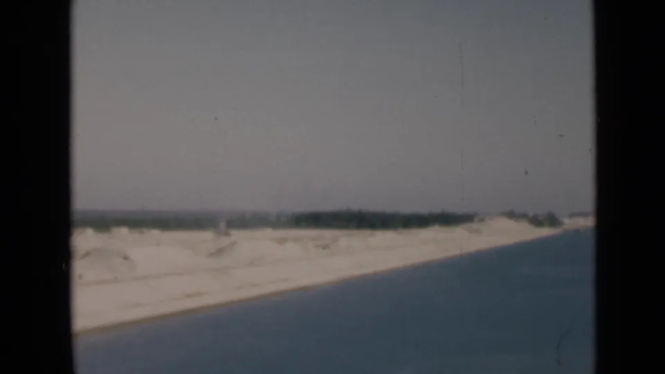1946 Suez Canal home-movie frame with blue water, white sand, distant green shoreline, dark borders, and visible film wear.