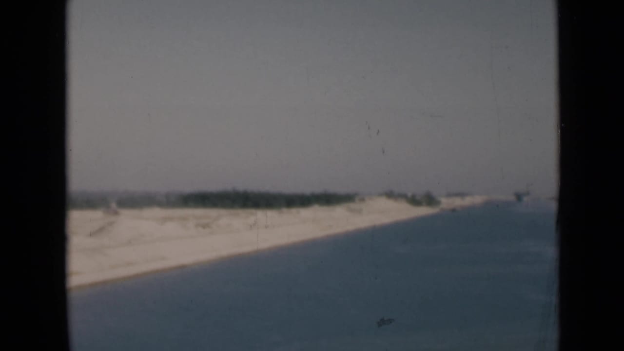 1946 Suez Canal home-movie frame showing calm blue water, pale sandy banks, and a faint distant shoreline.