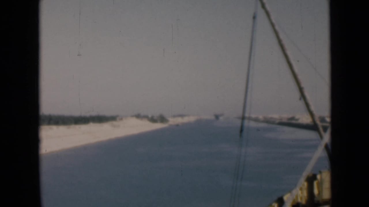 1946 Suez Canal view from a moving boat, with blue water, pale shoreline, distant vegetation, and rigging at the right edge.