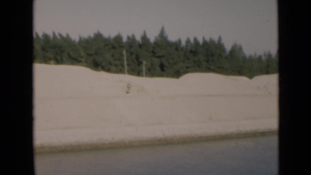 Suez Canal shoreline in 1946, showing a pale sandy bank, calm water and a dark tree line beyond the embankment.