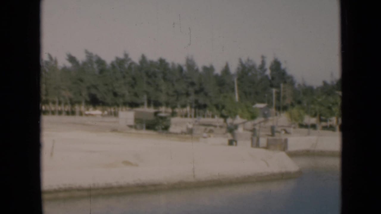 1946 Suez Canal bank with small shoreline structures, posts, trees and water visible from a passing boat.
