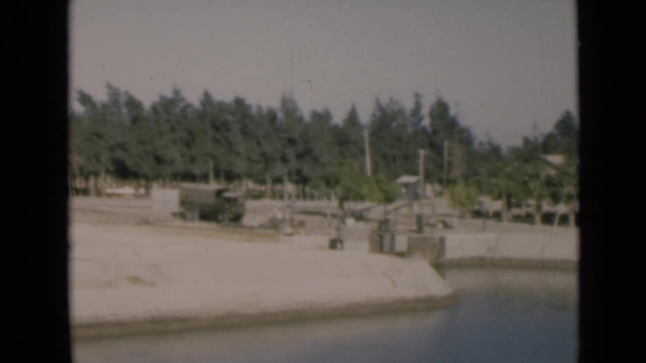 Canal water beside a concrete embankment with trees, poles, and a dark truck in the distance.