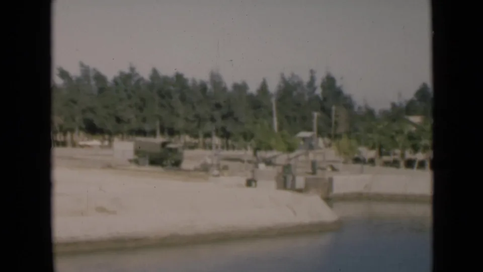 1946 Suez Canal home-movie frame showing calm water, a concrete bank, roadside trees, utility poles, and a dark truck beyond the canal edge.