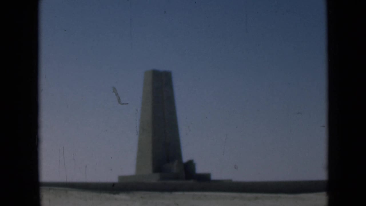 Tall obelisk-like monument against blue sky, framed by dark 8mm borders.