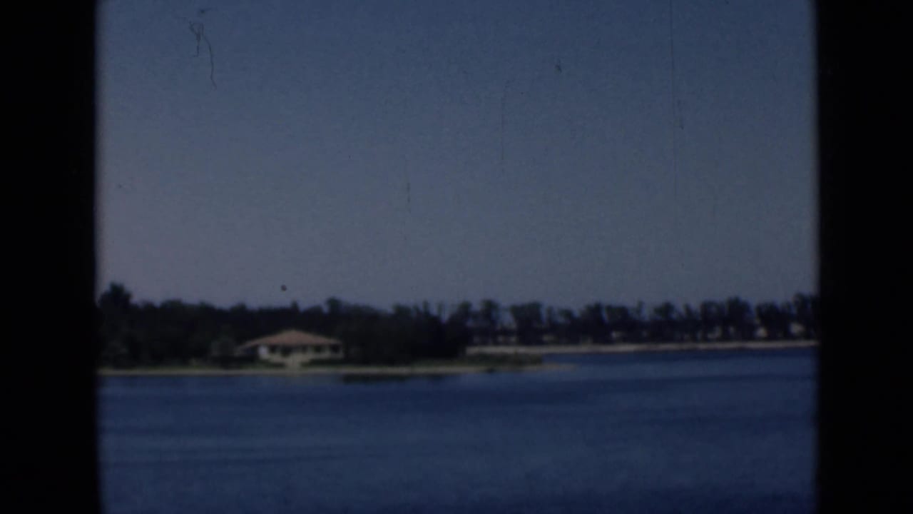 1946 8mm view of the Suez Canal area with calm water, a low shoreline, trees, and a small building in bright daylight.