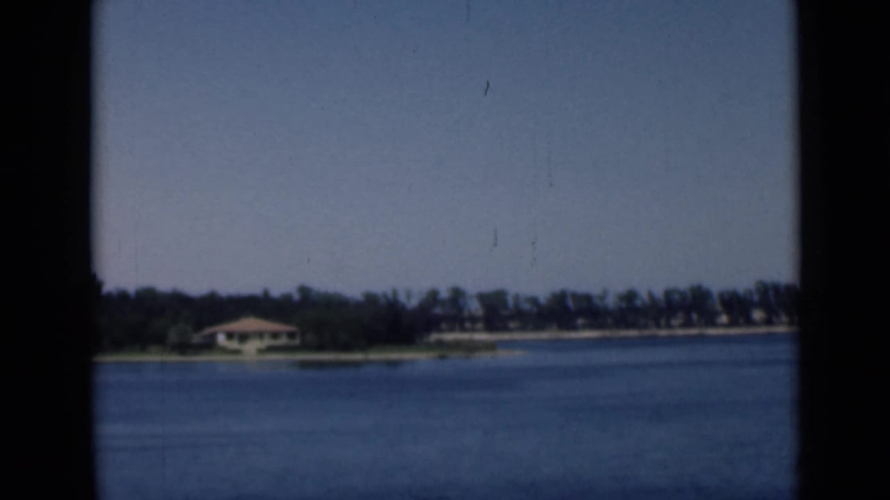 1946 Suez Canal home-movie frame showing blue water, a far tree line, and low shoreline buildings under a clear sky.