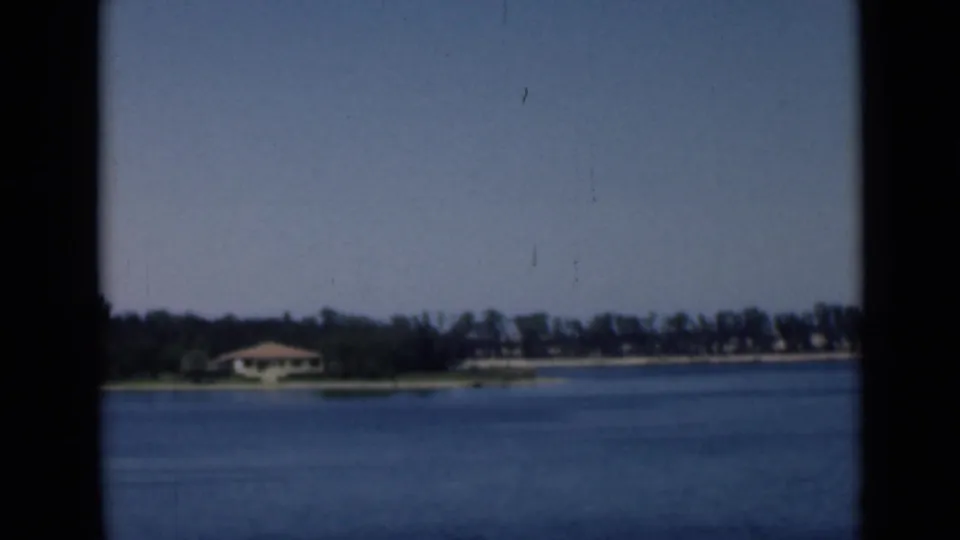 1946 Suez Canal home-movie frame showing blue water, a far tree line, and low shoreline buildings under a clear sky.