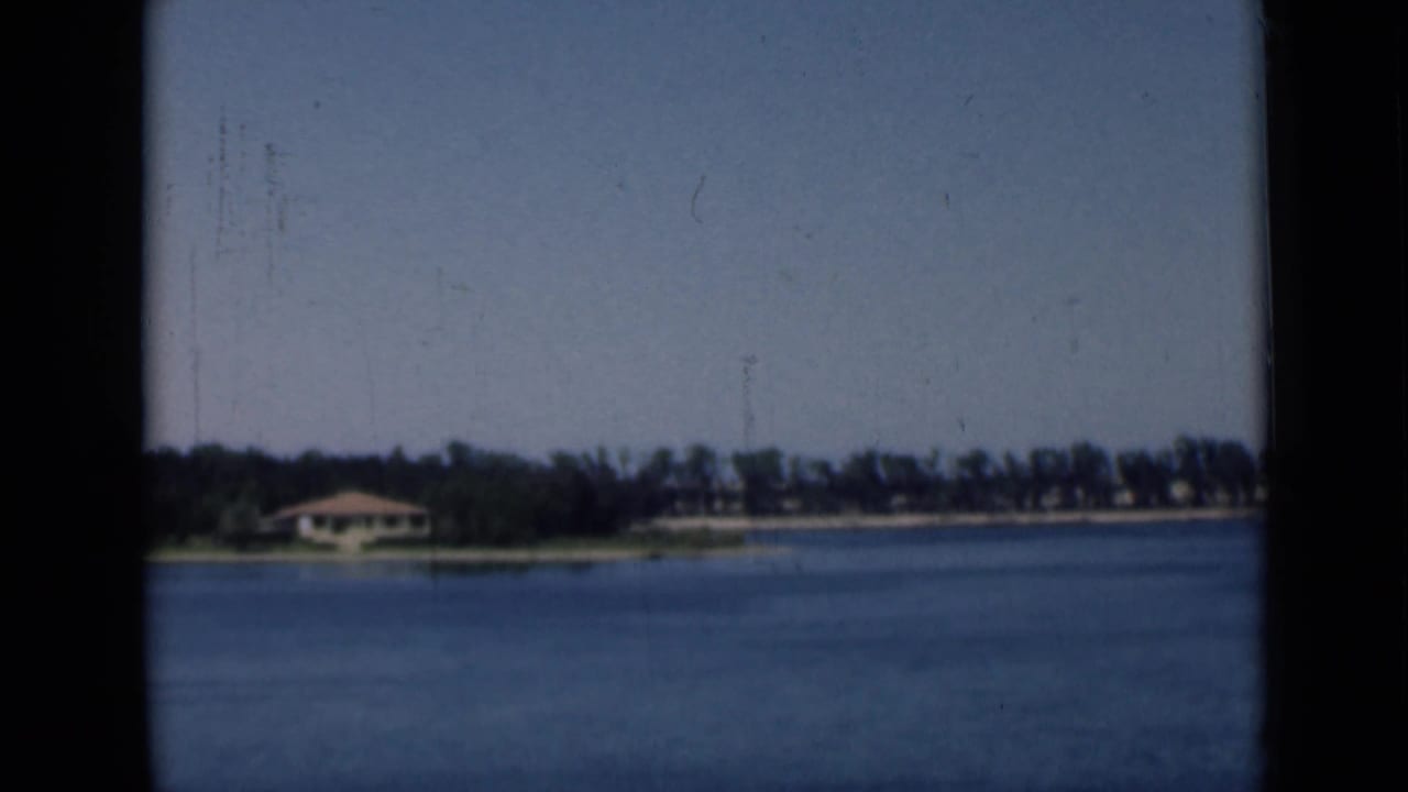 1946 8mm canal-side view in Egypt with a small shoreline building, trees, calm blue water, and a pale open sky.