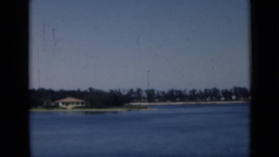 1946 8mm canal-side view in Egypt with a small shoreline building, trees, calm blue water, and a pale open sky.