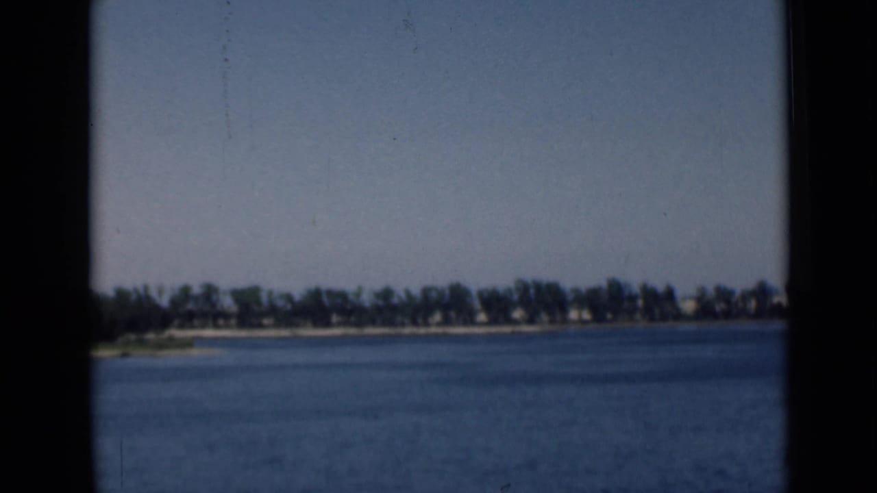 1946 Suez Canal frame with broad blue water, hazy pale sky, distant tree line, and low buildings along the far shore.