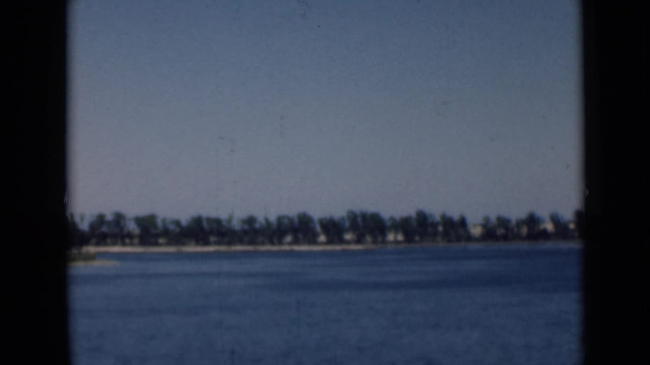 1946 Suez Canal home-movie still showing open water, a far tree-lined bank, small shoreline buildings, and visible film grain.