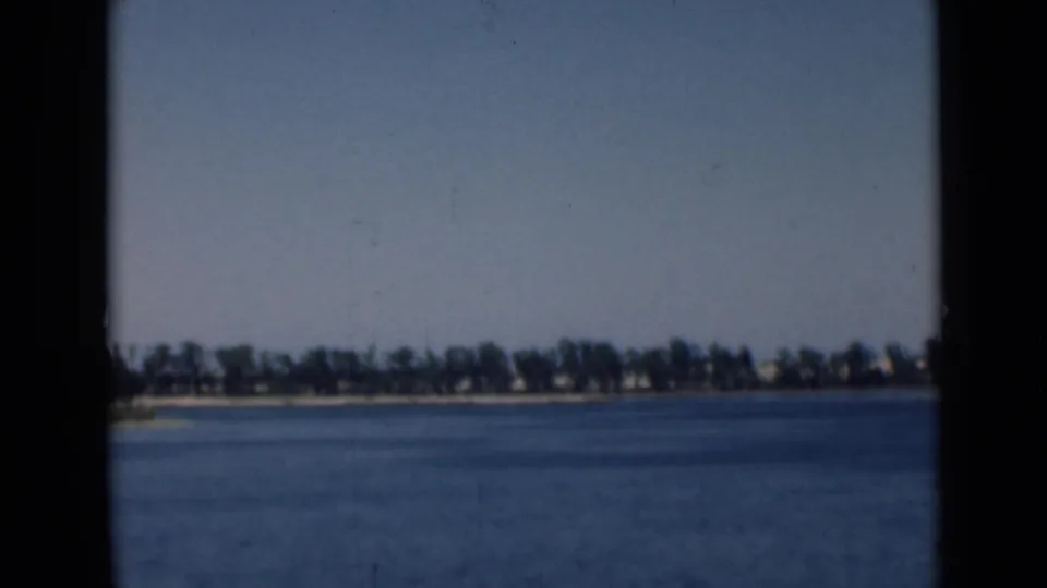 1946 Suez Canal home-movie still showing open water, a far tree-lined bank, small shoreline buildings, and visible film grain.