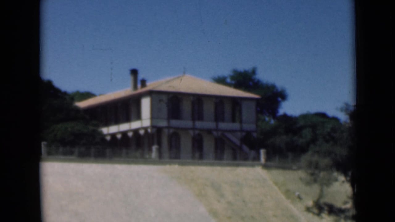 1946 Suez Canal area home-movie frame showing a two-story house with pale walls, tiled roof, veranda, trees, and dry foreground grass.