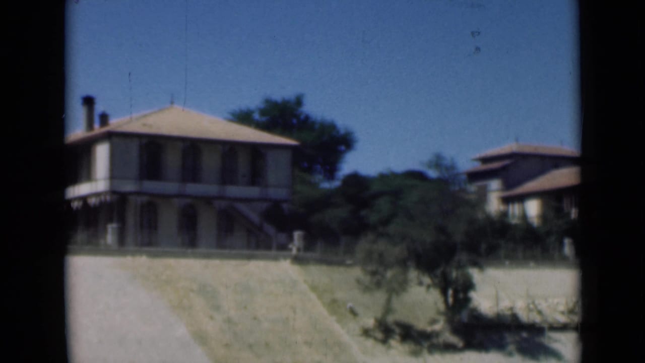 1946 Suez Canal area frame with a pale two-story house, a second house at right, trees, blue sky, and sunlit dry ground.