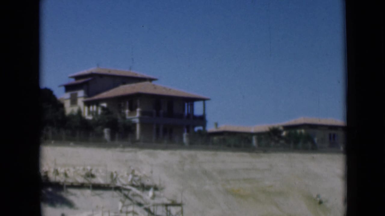 Shorefront buildings near the Suez Canal area appear across pale sand, framed by dark 8mm gate borders and blue sky.