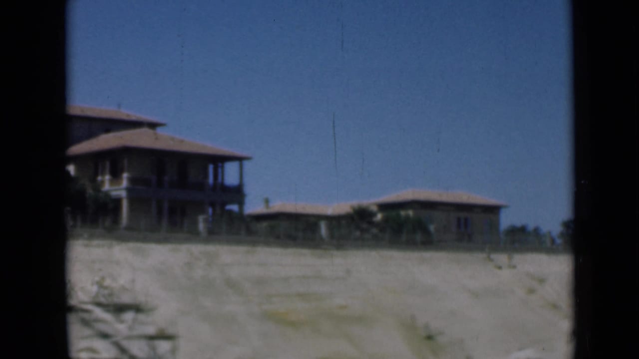 A 1940s home-movie view shows sandy shoreline terrain with a large house partly at left and a low building farther back.