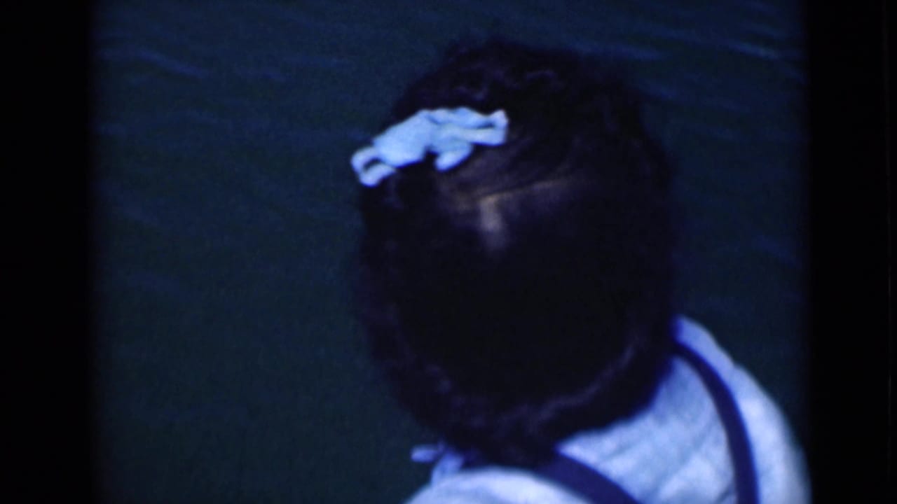 Brief close view of a woman in a light top, her dark hair marked by a pale blue accessory, against a dark green background.