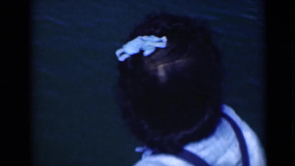 Brief close view of a woman in a light top, her dark hair marked by a pale blue accessory, against a dark green background.