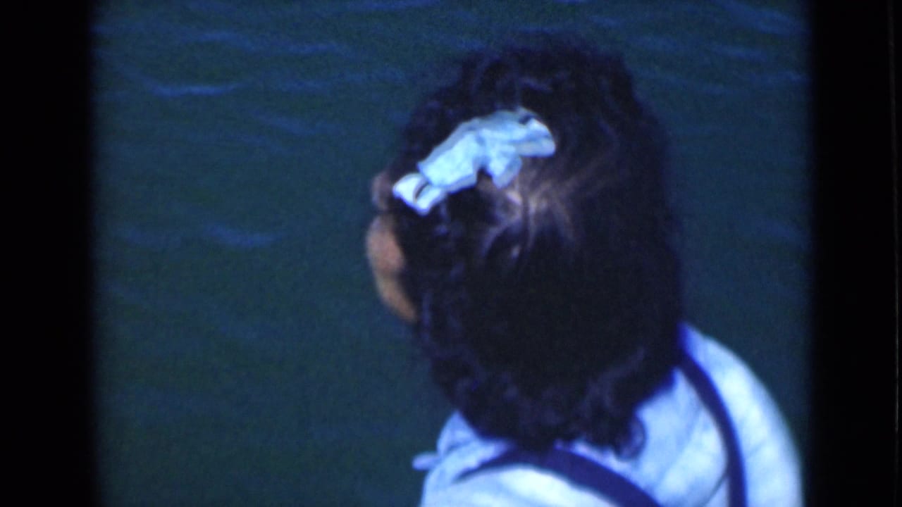 The woman turns away from the camera, showing her blue hair bow and light top against a water backdrop.