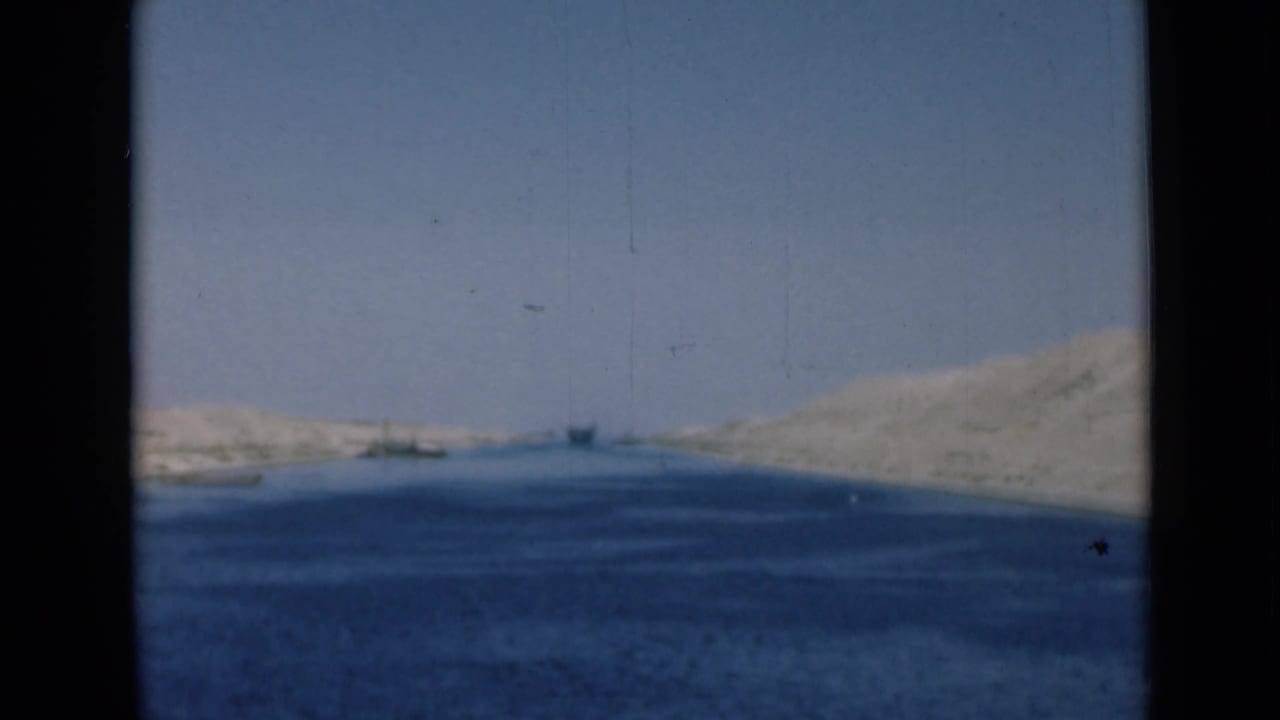 Wide view along the Suez Canal showing blue water between sandy banks, with small distant boats under a clear sky.