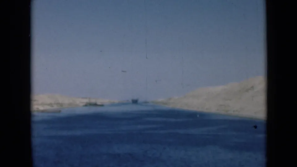 Wide view along the Suez Canal showing blue water between sandy banks, with small distant boats under a clear sky.
