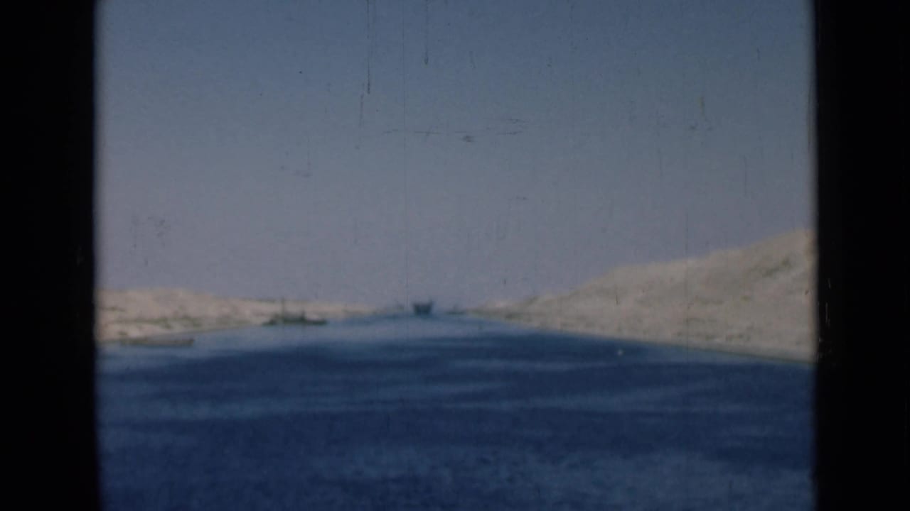 Suez Canal landscape with calm blue water, pale sandy banks, and small distant vessels in a 1940s home-movie frame.