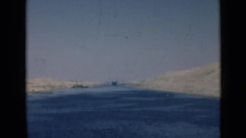 Suez Canal landscape with calm blue water, pale sandy banks, and small distant vessels in a 1940s home-movie frame.