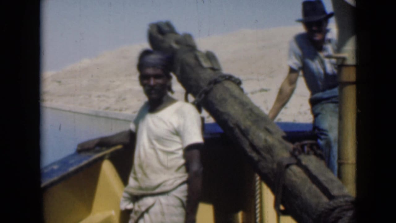 A man stands aboard a yellow and blue boat on the Suez Canal, with a large wooden spar and sandy bank behind him.
