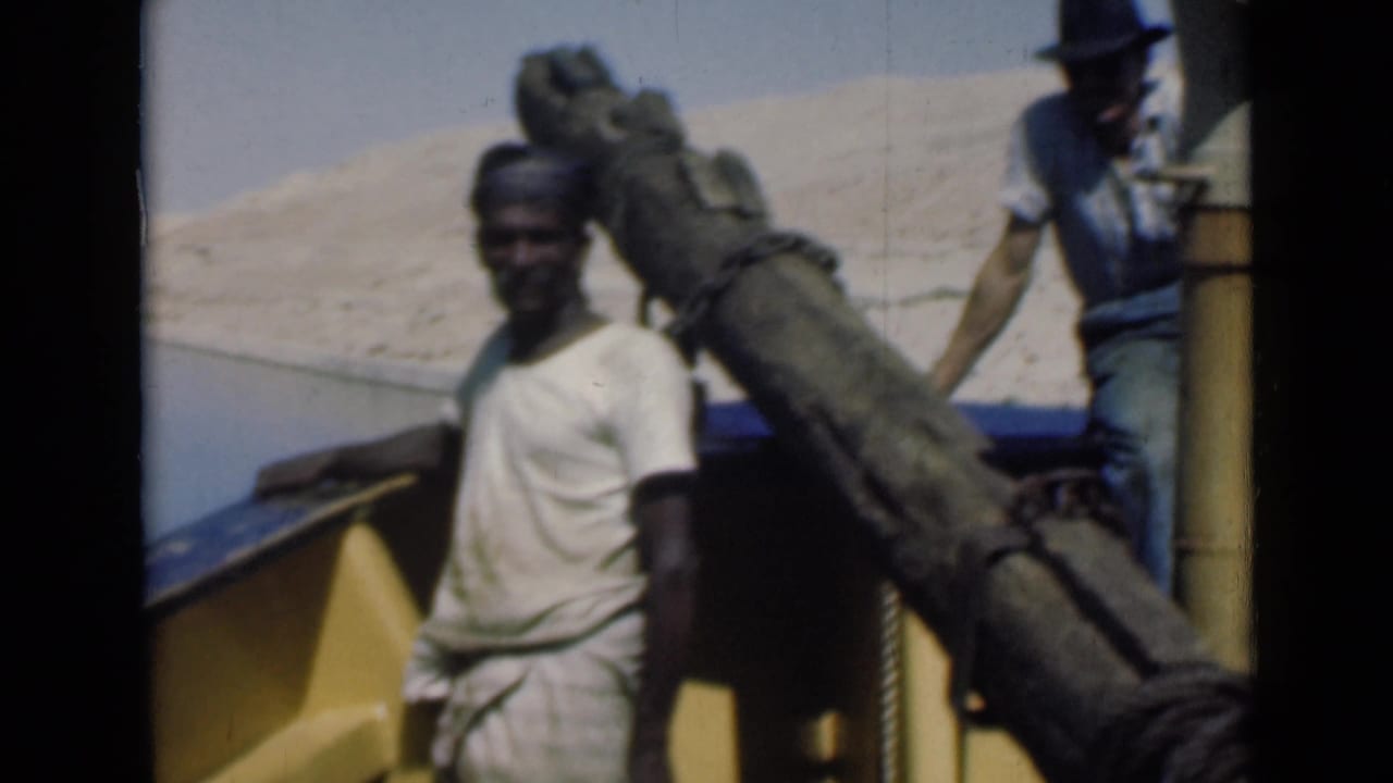 A worker in light clothing stands on the Suez Canal boat near rope and a large wooden spar, with desert shoreline in the background.