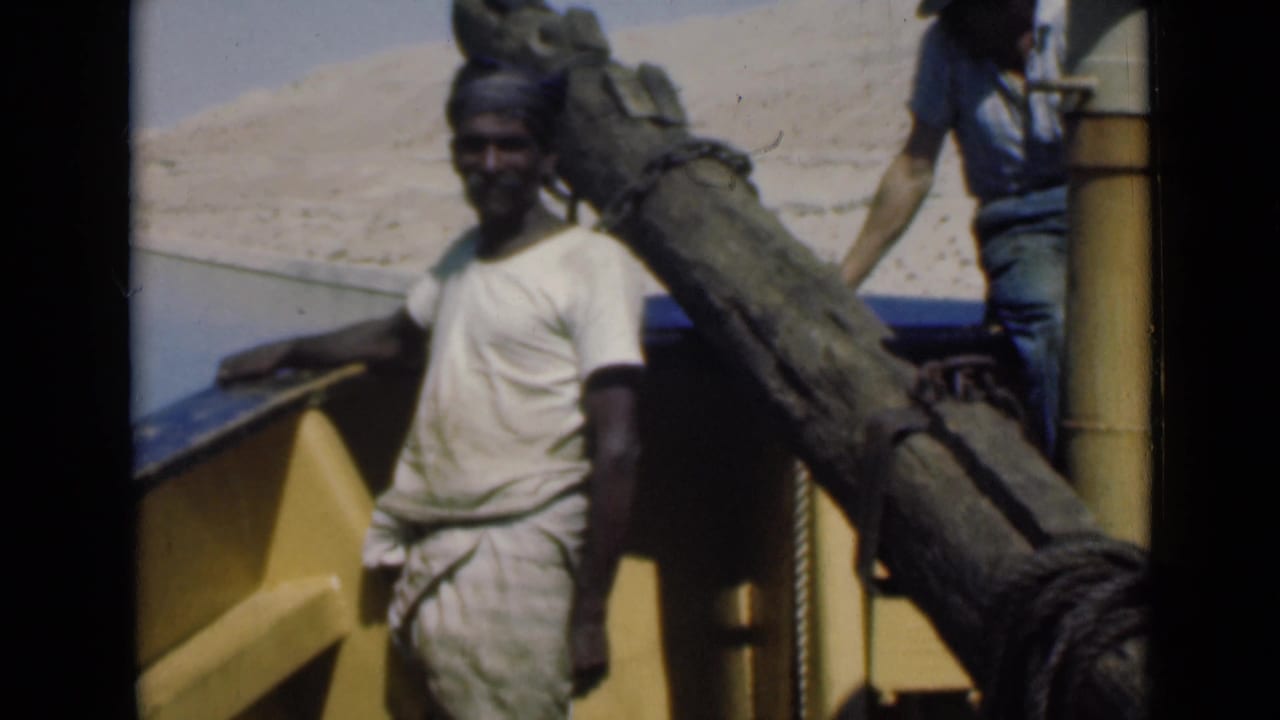 A man stands beside a large wooden spar on the Suez Canal boat, with another figure partly visible near a vertical post.