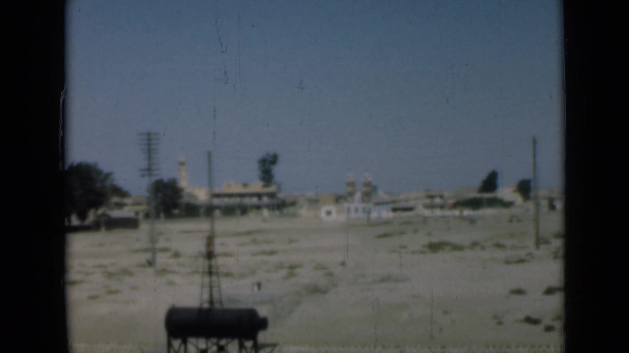 1946 Suez Canal area frame showing a distant settlement with low buildings, utility poles, open sandy ground, and a minaret-like tower.