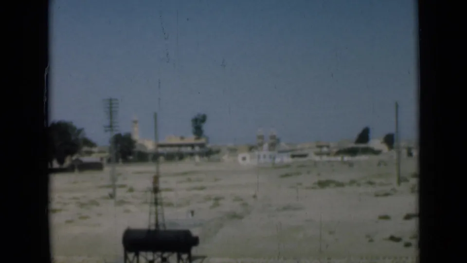 1946 Suez Canal area frame showing a distant settlement with low buildings, utility poles, open sandy ground, and a minaret-like tower.