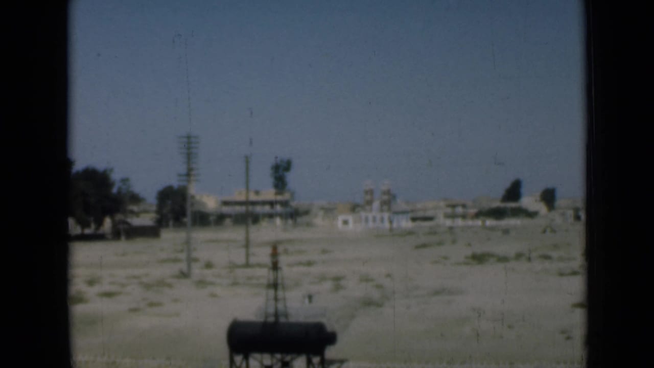1946 Suez Canal-area home-movie frame showing sandy open ground, utility poles, distant buildings, trees, and a dark cylindrical tank.
