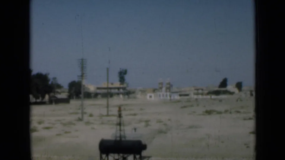 1946 Suez Canal-area home-movie frame showing sandy open ground, utility poles, distant buildings, trees, and a dark cylindrical tank.