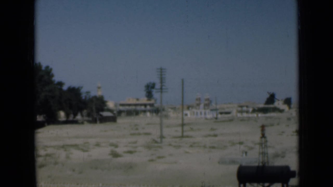 1946 Suez Canal-area frame with open sandy terrain, utility poles, low buildings, a mosque-like structure, trees, and a dark tank.