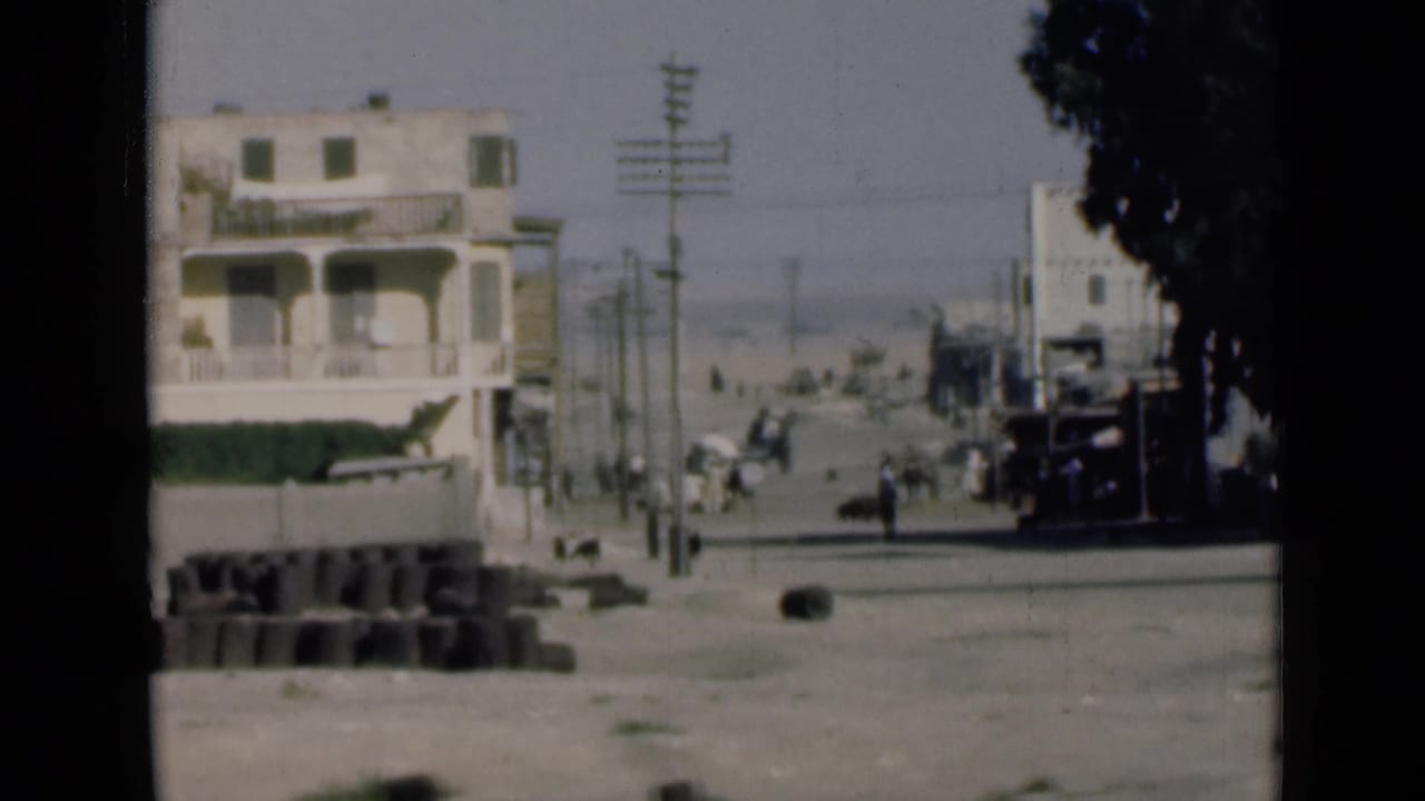 1946 8mm frame from near the Suez Canal showing a pale building, roadside blocks, utility poles, pedestrians, and a broad dusty road.
