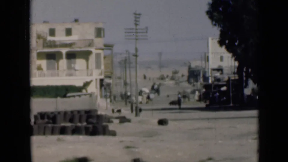 1946 8mm frame from near the Suez Canal showing a pale building, roadside blocks, utility poles, pedestrians, and a broad dusty road.