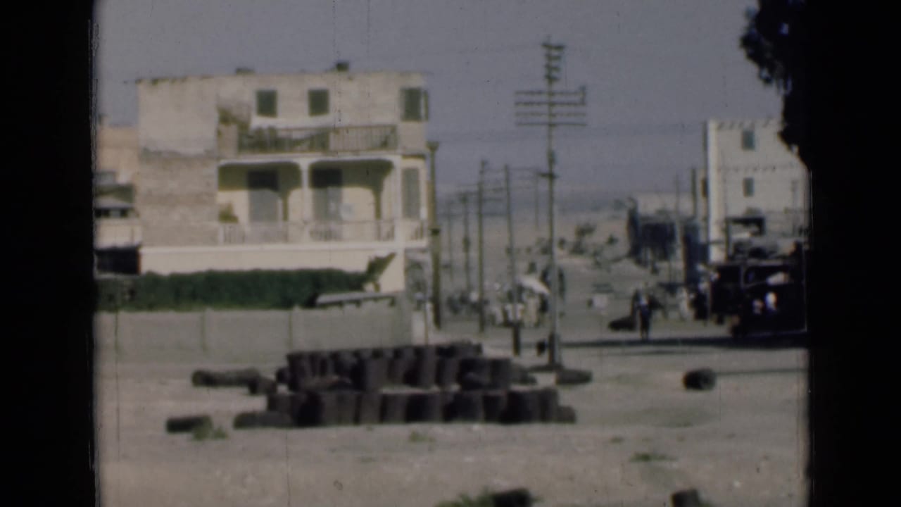1946 Egypt home-movie frame near the Suez Canal with a pale building, roadside blocks, utility poles, pedestrians, trees, and vehicles.